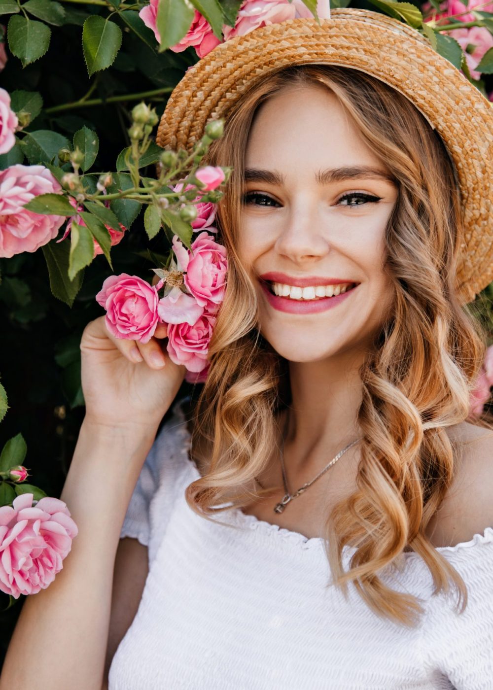 Adorable girl with curly blonde hair posing in garden. Portrait of caucasian glad woman holding pink flower.