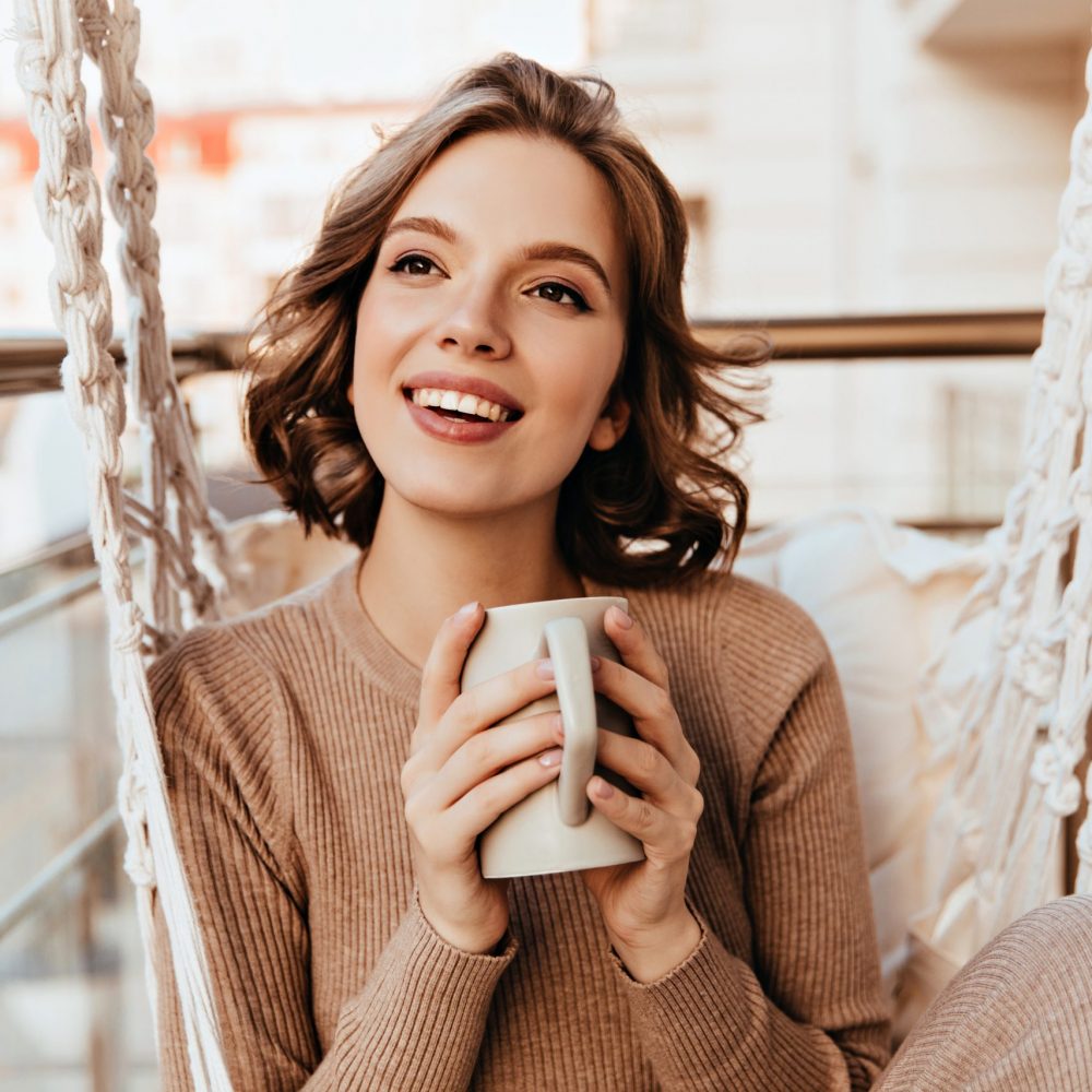 Carefree girl with brown makeup drinking tea at balcony. Photo of pleasant brunette woman in knitted dress enjoying coffee.